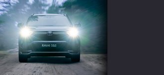 TOYOTA RAV4 Plugin Hybrid Direkte Frontalansicht der Vorderseite eines TOYOTA RAV4 Plugin Hybrid mit schwarzem Lack. Der Neuwagen kommt mit leuchtenden Scheinwerfern auf die Kamera zu uns fährt durch ein kleines Waldstück. Ein drittel des Bildes wird mit einem grauen Balken überdeckt.