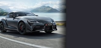 TOYOTA SUPRA Schrägansicht der Vorderseite eines TOYOTA SUPRA mit grauem Lack. Der Neuwagen fährt auf einer Straße. Im Hintergrund ist ein See mit einer schönen Bergsilhouette zu sehen. Ein drittel des Bildes wird mit einem grauen Balken überdeckt.