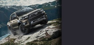 TOYOTA HILUX Schrägansicht auf die Vorderseite eines TOYOTA HILUX mit dunkelgrauem Lack. Der Neuwagen steht an einem Berghang und fährt den unebenen Boden nach oben. Im Hintergrund ist eine Bergsilhouette zu sehen. Ein drittel des Bildes wird mit einem grauen Balken überdeckt.