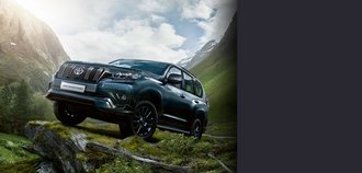 TOYOTA LAND CRUISER Schrägansicht auf die Vorderseite eines TOYOTA LAND CRUISER mit dunkelblauem, fast grauem Lack. Der Neuwagen steht auf einem Felsen an einem Berghang. Im Hintergrund sind Gipfel von hohen Bergen zu sehen. Ein drittel des Bildes wird mit einem grauen Balken überdeckt.