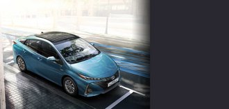 TOYOTA PRIUS Plugin Hybrid Erhöhte Schrägansicht auf die Vorderseite eines TOYOTA PRIUS Plugin Hybrid mit himmelblauem Lack. Der Neuwagen wurde auf einem Parkplatz in der Stadt am hellichten Tag geparkt. Ein drittel des Bildes wird mit einem grauen Balken überdeckt.