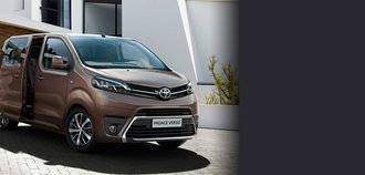 TOYOTA PROACE City Verso Schrägansicht auf die Vorderseite eines TOYOTA PROACE City Verso mit braunem Lack. Der Familienvan ist vor einer Garage eines Hauses geparkt und steht dort mit offener Tür. Ein drittel des Bildes wird mit einem grauen Balken überdeckt.