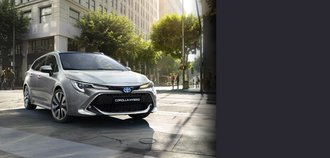 TOYOTA COROLLA Touring Sports Schrägansicht auf die Vorderseite eines TOYOTA COROLLA Touring Sports mit silbernem Lack. Der Neuwagen steht auf einem hellen Stadtplatz und wird von Tageslicht beleuchtet. Ein drittel des Bildes wird mit einem grauen Balken überdeckt.